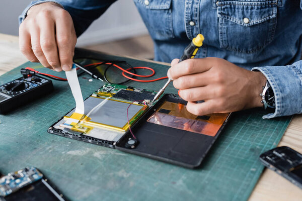 Close up view of hands of repairman with flex cable and screwdriver near disassembled digital tablet with multimeter on workplace