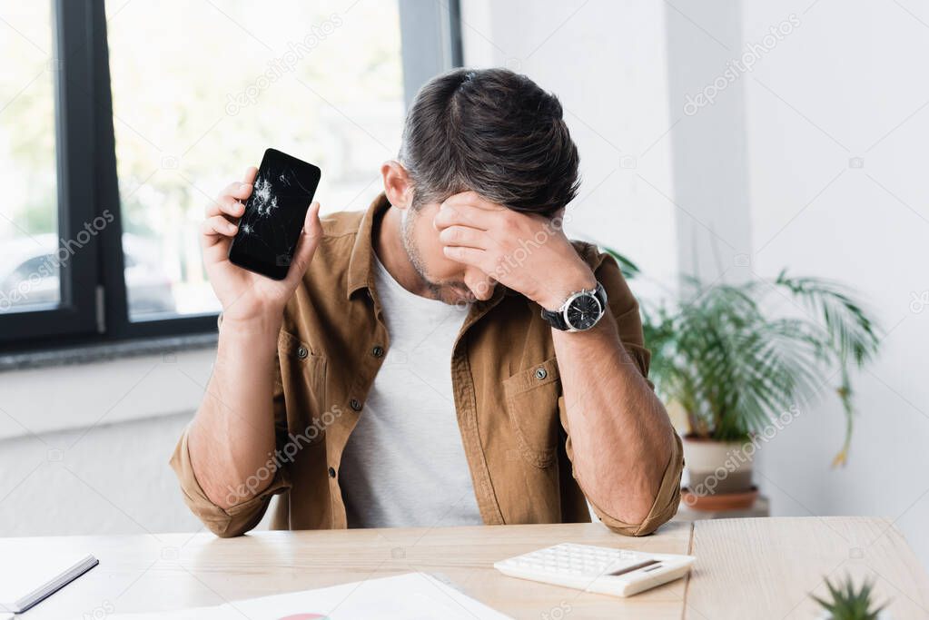 Disappointed businessman with smashed smartphone sitting at workplace on blurred background