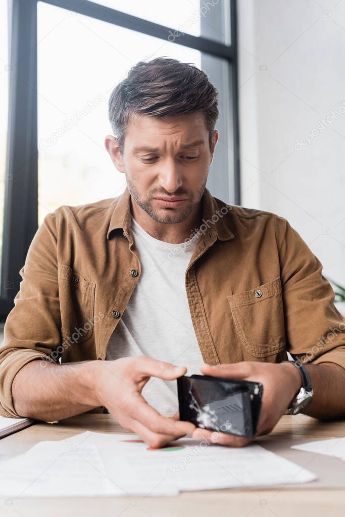 Sad businessman disassembling damaged smartphone while sitting at workplace on blurred background