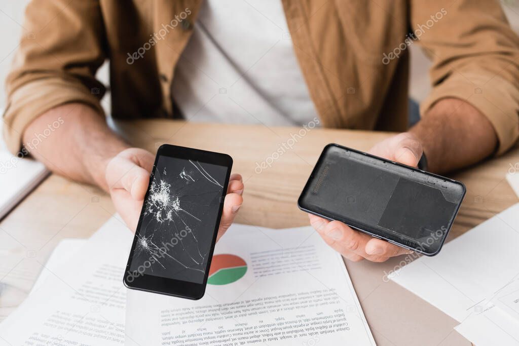 Cropped view of businessman showing disassembled smashed smartphone at workplace on blurred background