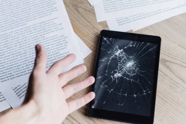 Cropped view of man with hand near smashed digital tablet and documents on wooden background clipart