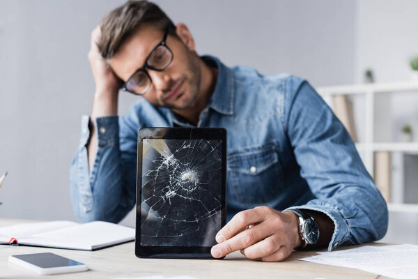 Sad businessman with smashed digital tablet sitting at workplace on blurred background