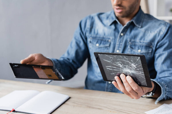 Cropped view of businessman disassembling digital tablet and holding smashed touchscreen at table on blurred background