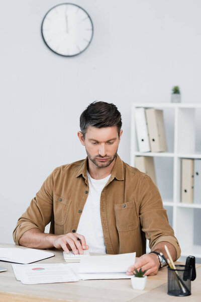 Focused businessman looking at paper sheet while counting on calculator at workplace on blurred background