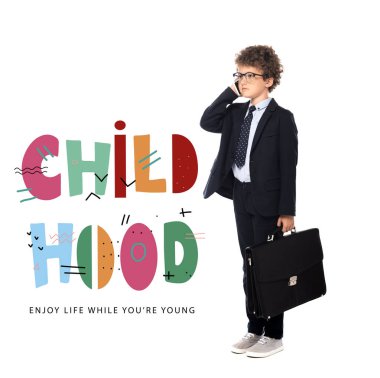 curly boy in suit and glasses holding briefcase while talking on smartphone near childhood, enjoy life while you are young lettering on white clipart