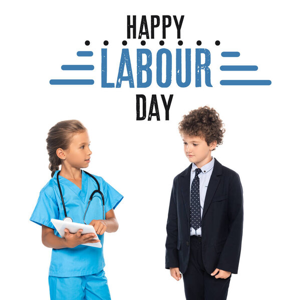 child in costume of doctor holding digital tablet near curly boy in formal wear and happy labour day lettering on white 