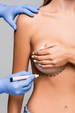 Kırpılmış plastik cerrah görüşü, gri üzerine izole edilmiş genç bir kadının göğsüne yakın bir işaret tutuyor. 