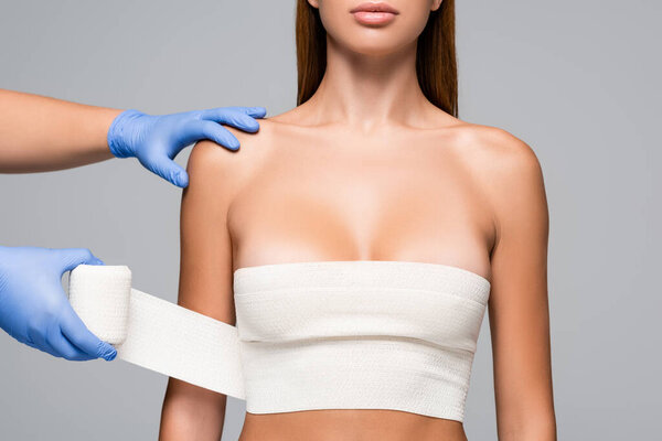 Cropped view of doctor in latex gloves wrapping breast of naked woman with elastic bandage isolated on grey 