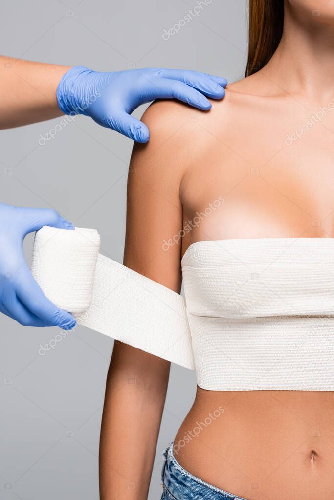 Cropped view of doctor wrapping breast of naked woman with elastic bandage isolated on grey 