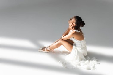 graceful african american ballerina in dress sitting on floor on white background clipart