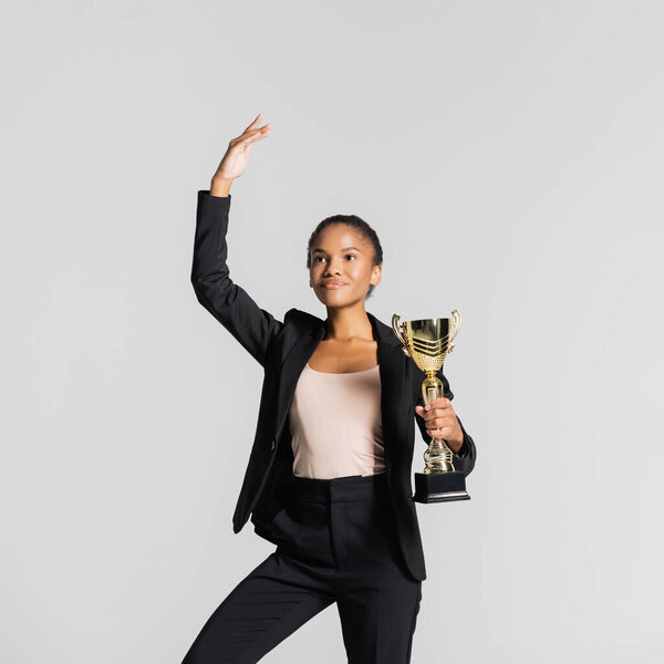smiling african american businesswoman performing ballet with golden cup isolated on grey