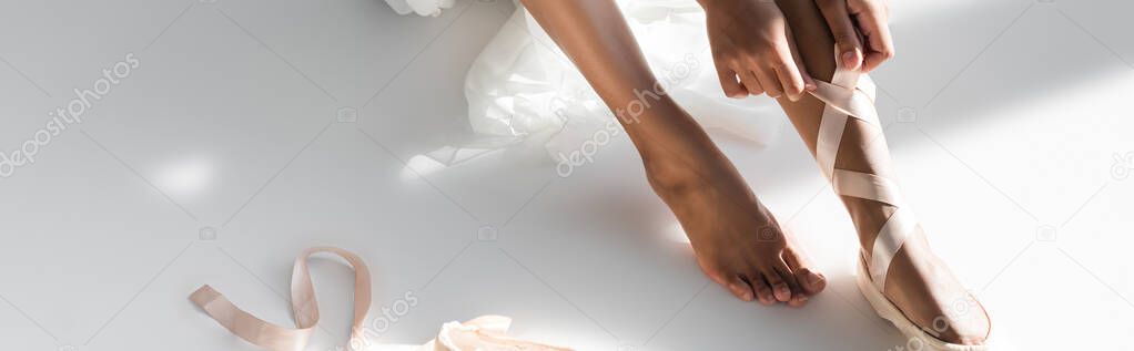 cropped view of graceful african american ballerina tying pointe shoes on white background, banner
