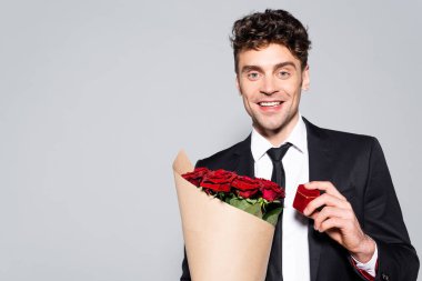 smiling man in elegant suit with bouquet of red roses and ring box isolated on grey clipart