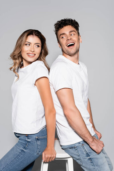smiling young couple in white t-shirts and jeans posing back to back isolated on grey