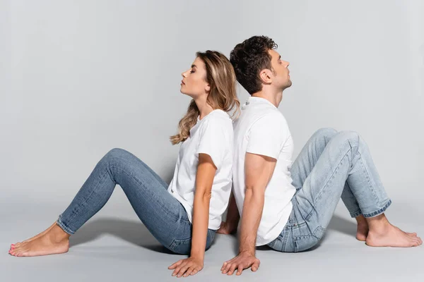 young couple in white t-shirts and jeans posing back to back isolated on grey