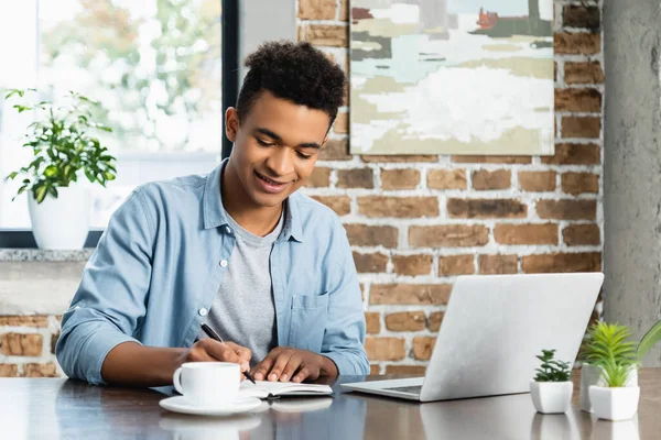 Hombre Afroamericano Feliz Escribiendo Cuaderno Cerca Computadora Portátil Taza Escritorio —  Fotos de Stock