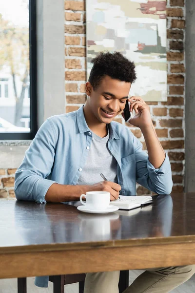 Hombre Afroamericano Feliz Hablando Teléfono Inteligente Escribiendo Cuaderno —  Fotos de Stock