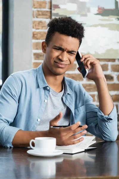 Homem Americano Africano Descontente Falando Smartphone Segurando Caneta Perto Notebook — Fotografia de Stock