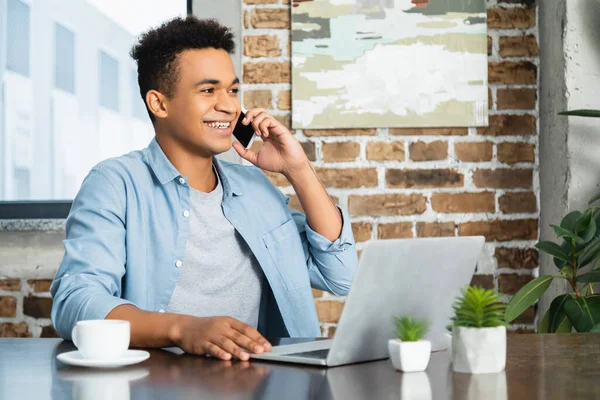 Sonriente Afroamericano Hombre Hablando Teléfono Inteligente Cerca Computadora Portátil Escritorio —  Fotos de Stock