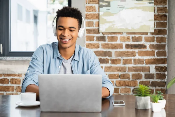 Kablosuz kulaklıklı mutlu Afrikalı Amerikalı adam masadaki dizüstü bilgisayarın yanında podcast dinliyor.