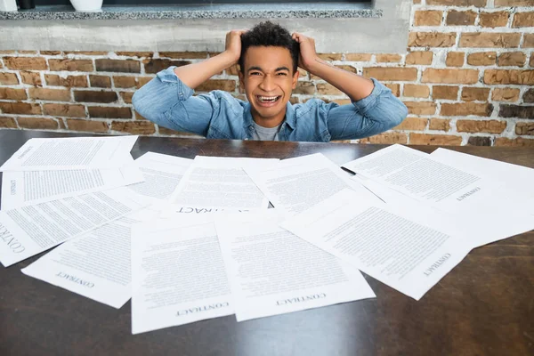 hysterical african american man pulling hair near documents with contract lettering on desk