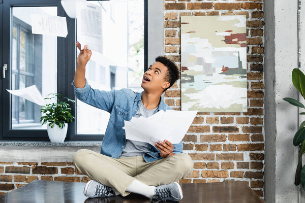 изумленный африканский американец, сидящий на столе и бросающий воздушные документы 