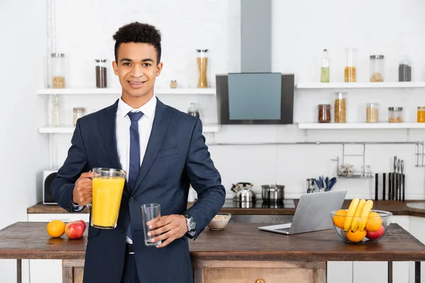 快乐的非洲商人拿着装有橙汁和杯子的罐子在厨房里 — 图库照片