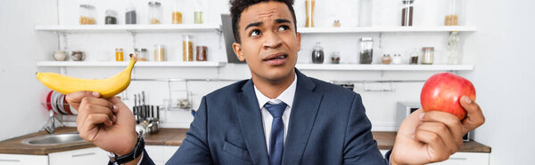 puzzled african american businessman choosing between apple and banana, banner