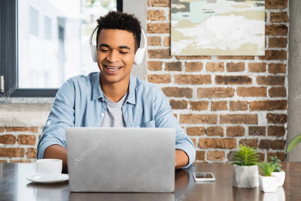 happy african american man in wireless headphone listening podcast near laptop on desk