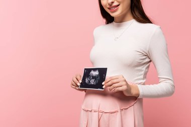 cropped view of happy young pretty pregnant woman holding ultrasound scan isolated on pink clipart
