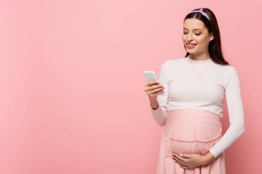 happy young pretty pregnant woman using smartphone isolated on pink clipart