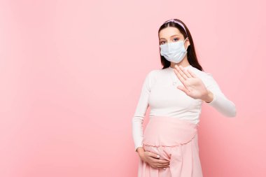 young pregnant woman in medical mask showing stop gesture isolated on pink clipart