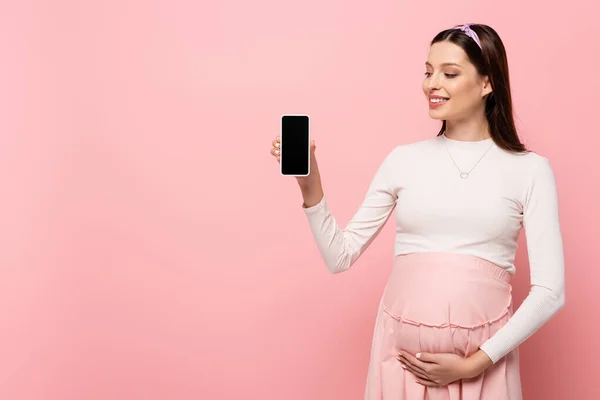 幸せな若い妊娠中の女性がピンクに隔離されたスマートフォンを見せ — ストック写真