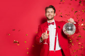 Stylový muž v obleku s úsměvem na kameru při držení disco koule a sklenice šampaňského na červeném pozadí 
