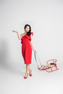 Cheerful woman in red dress and heels holding sleigh on grey background clipart