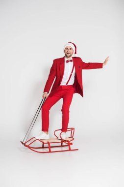 Smiling man in santa hat and red suit looking at camera while standing on sleigh on grey background  clipart