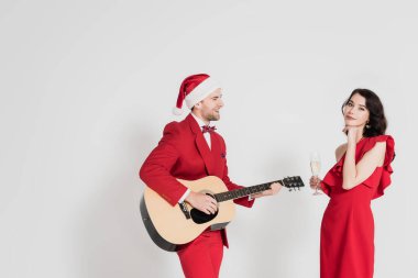 Saten şapkalı gülümseyen adam gri arka planda bir bardak şampanyası olan kadının yanında akustik gitar çalıyor. 