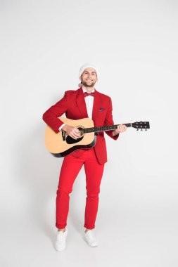 Positive man in suit and santa hat playing acoustic guitar on grey background  clipart