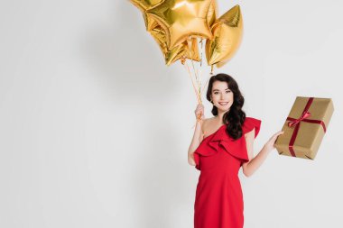 Elinde balonlar ve gri arka planda hediye paketi tutan gülümseyen kadın.