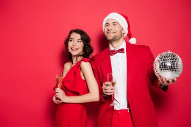 Şampanya kadehleri, disko topu ve kırmızı arka planda Noel Baba şapkası olan gülümseyen çift. 