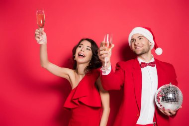Disko topu ve Noel Baba şapkalı neşeli çift kırmızı arka planda şampanya bardakları tutuyor. 