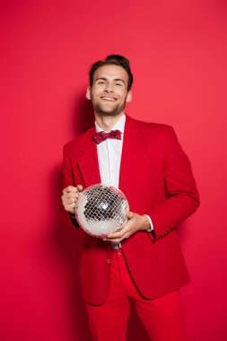 Kırmızı elbiseli gülümseyen adam kırmızı arka planda disko topu tutuyor. 