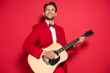 Kırmızı arka planda akustik gitar çalan takım elbiseli neşeli bir adam. 