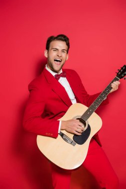 Kırmızı arka planda akustik gitar çalan takım elbiseli neşeli bir adam. 