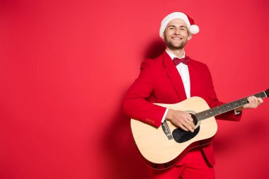 Smiling man in santa hat playing acoustic guitar on red background  clipart