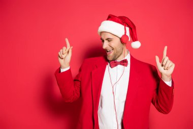 Noel Baba şapkalı heyecanlı adam kulaklıkla müzik dinliyor ve kırmızı arka planı işaret ediyor. 