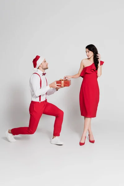 男でサンタ帽子保持ギフトの間に1膝の近くにガールフレンドで赤いドレス上の灰色の背景 — ストック写真