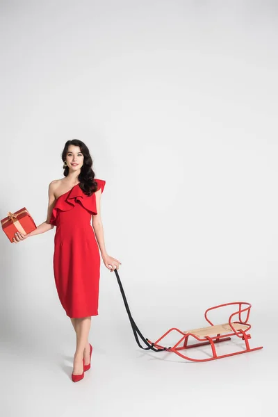 Brunette Woman Red Dress Heels Holding Present Sleigh Grey Background — Stock Photo, Image