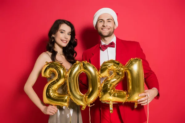 笑着戴大礼帽的男人拿着2021号形状的气球 身边的女朋友穿着红色背景的衣服 — 图库照片