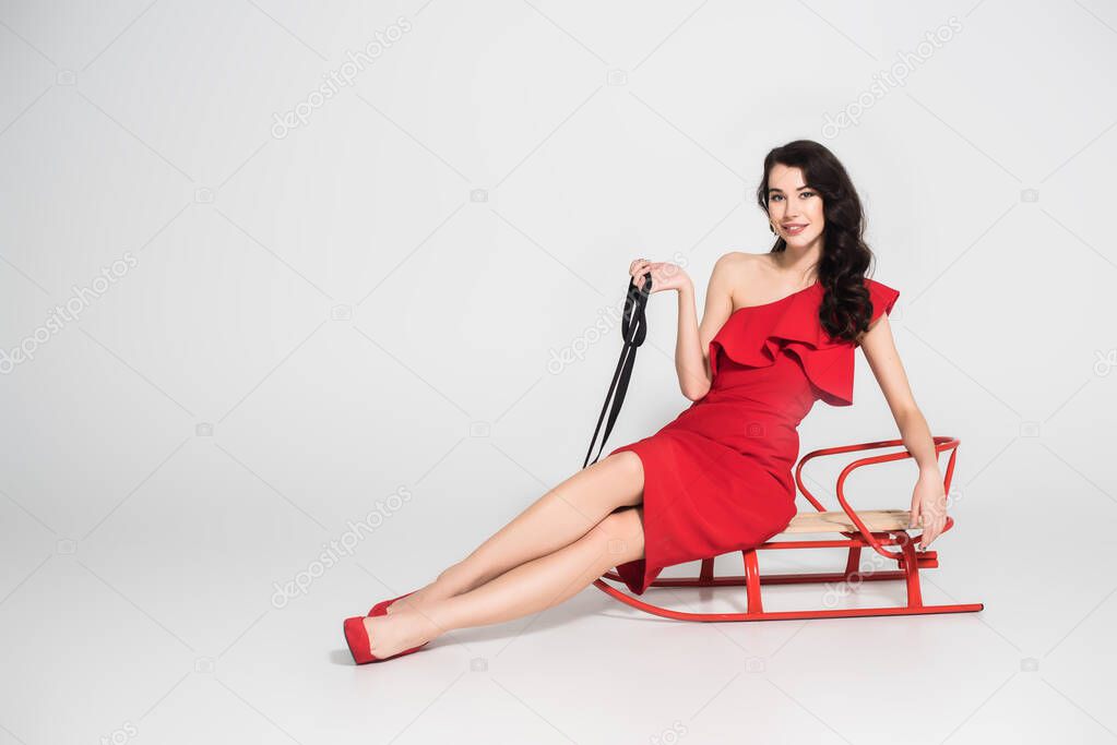 Brunette woman in red dress smiling at camera while sitting on sleigh on grey background